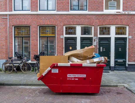 A large red waste skip positioned on the pavement in front of three dark green front doors of a brick building, situated on a residential street in Southfields. The skip is filled with various discarded items including flattened cardboard boxes, plastic packaging, and miscellaneous rubbish, indicating recent rubbish removal activity. To the left of the skip, two bicycles are parked leaning against the building wall, with one bike fitted with a black basket on its handlebars. The building features tall, narrow windows with metal grilles and decorative yellow accents above the windows, typical of period residential properties in Southfields. The overall scene is illuminated by daylight, with a slightly overcast sky, creating a neutral, natural lighting environment suitable for an office clearance or waste collection service. This image visually supports waste management activities conducted by Office Clearance Southfields, demonstrating rubbish removal in an urban residential setting within the SW19 postcode area.