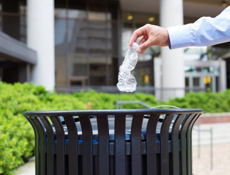 A person's hand, dressed in a light blue shirt, is seen dropping a crumpled clear plastic bottle into a black slatted outdoor rubbish bin, which is positioned on a paved surface with a landscaped green area in the background. The scene appears to be outside a modern office or commercial building, with glass windows and structural elements visible in the background. The image emphasizes waste disposal and rubbish collection, relevant to the rubbish removal services provided by Office Clearance Southfields, located near Southfields and the Wandsworth area postcode. The focus is on responsible waste management, with natural daylight illuminating the scene and highlighting the contrast between the transparent bottle and the dark, sturdy bin. This visual reinforces the importance of proper rubbish disposal in urban and commercial environments associated with Southfields.