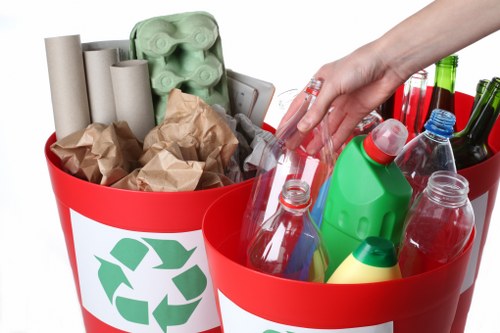 A pair of bright red plastic recycling bins filled with various waste materials, including crumpled brown paper, empty glass bottles in different colors such as green, clear, and brown, and cardboard tubes, all placed on a white background. The bin on the left contains brown paper packaging and cardboard tubes, while the bin on the right holds glass bottles and plastic containers with caps. A human hand, partially visible, is reaching into the bin on the right. The bins feature a green recycling symbol on the front, indicating their use for waste collection and rubbish disposal, in a context consistent with professional rubbish removal services in Southfields or nearby areas. The environment appears to be a controlled indoor setting, emphasizing waste segregation and recycling practices.
