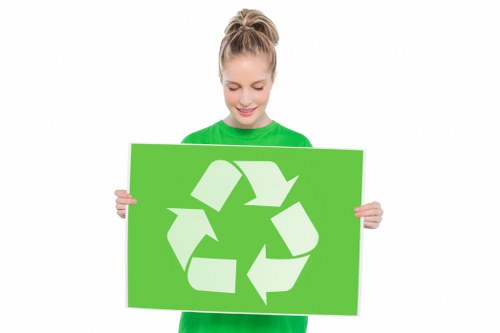 A young woman wearing a bright green shirt is holding a large rectangular green sign featuring a white recycling symbol composed of three arrows forming a triangle. She is smiling gently while standing against a plain white background. The recycling sign symbolizes environmentally responsible waste management, aligning with rubbish removal services offered by companies such as Office Clearance Southfields. The individual's neat appearance and the vibrant color scheme emphasize a positive message about recycling and waste reduction. This visual connects to eco-friendly rubbish disposal practices, particularly in urban areas like Southfields, suggesting a focus on responsible clearance and waste management solutions within the local community. The setting appears to be a studio or professional environment aimed at promoting sustainable waste practices, supporting services provided in the Southfields area, with a specific emphasis on rubbish removal from residential or commercial premises.