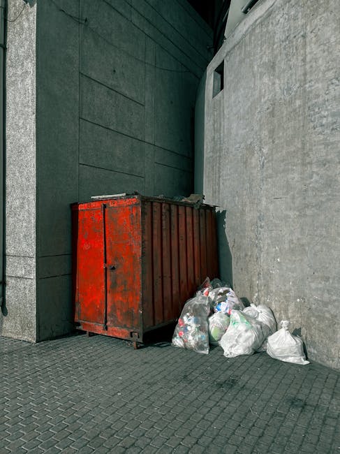 A large, weathered red metal skip with visible rust and chipped paint is positioned against a dark grey building wall on a paved urban footpath. Next to the skip, several tightly tied white and transp