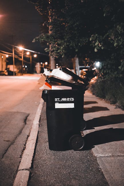 A waste collection worker dressed in a high-visibility orange and grey uniform is seen handling rubbish at the rear of a commercial waste collection vehicle during evening hours. The vehicle, which ha