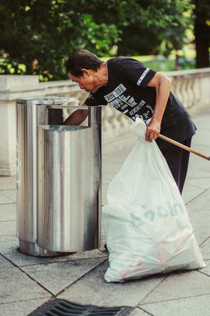 A worker dressed in green uniform and safety helmet is standing at the rear of a small waste collection vehicle, which has an open, dirty metal hopper section used for collecting rubbish. The vehicle