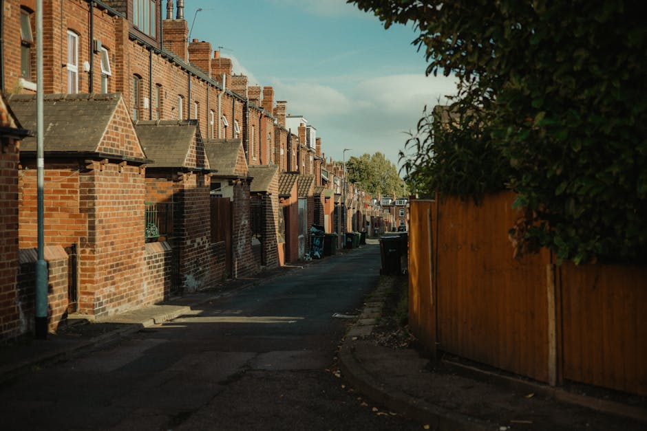 The image depicts a quiet residential street lined with Victorian-style terraced houses featuring white facades, sash windows, and tiled roofs with red chimneys. The foreground shows an empty asphalt