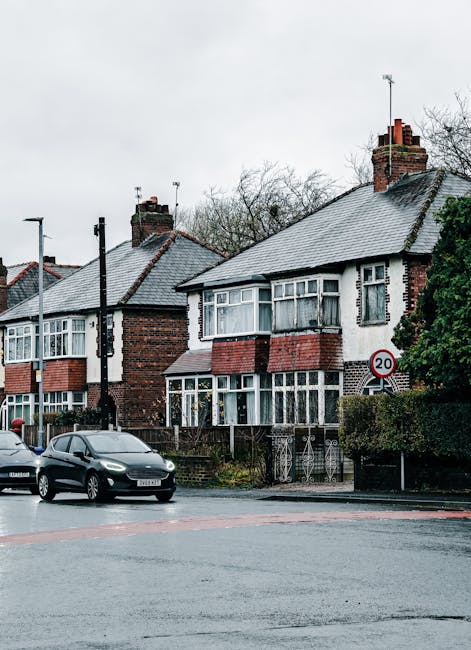 A quiet residential street scene featuring a row of Victorian-style terraced houses with ornate white facades, bay windows, and red brickwork, situated along a paved sidewalk. The houses are separated